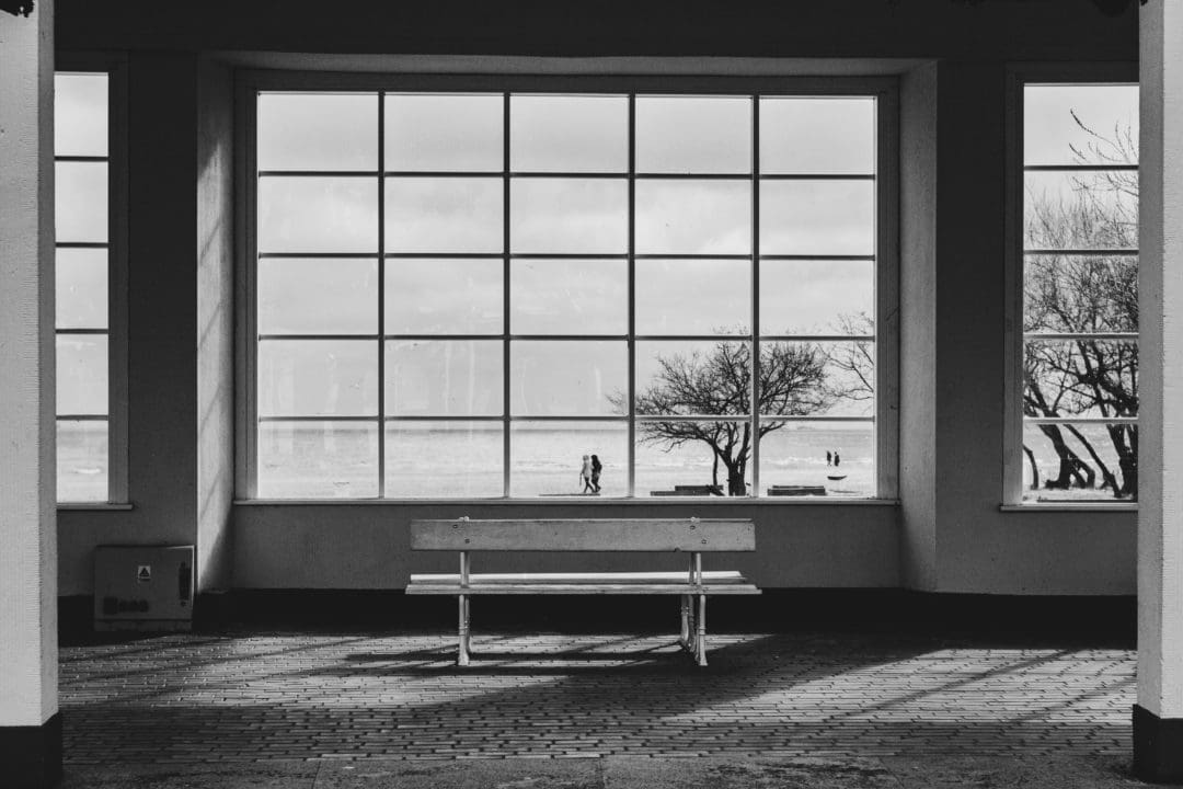 Eine Bank steht in einem Rundgang, dahinter ein großes Fenster mit Sicht auf Menschen am Strand