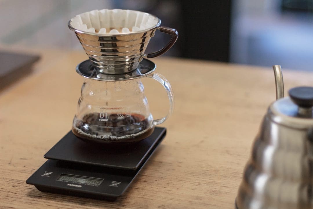 Eine Glaskanne mit Metall-Filter steht auf einer schwarzen Waage, der Kaffee tropft durch den Filter in die Kanne