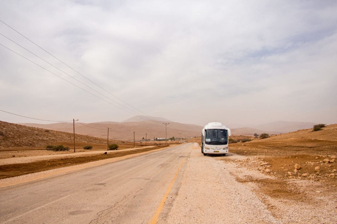 An einer staubigen kleinen Asphaltstraße steht ein weißer Bus, links verlaufen Strommasten, im Hintergrund ist eine hügelige Wüstenlandschaft zu sehen