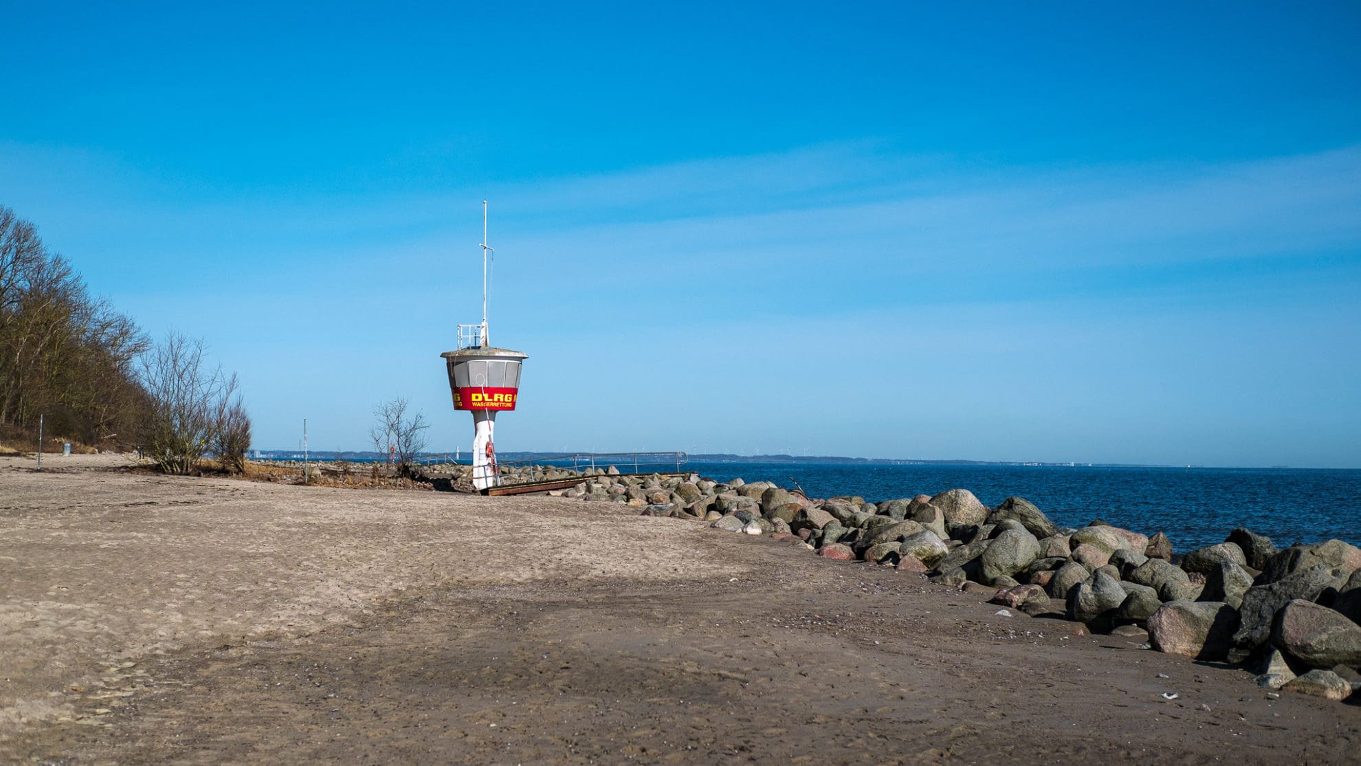 Ein Turm der DLRG mit weißem Fuß steht am Strand. Große Steine grenzen den Sandstrand vom Wasser ab. Der Himmel ist strahlend blau.