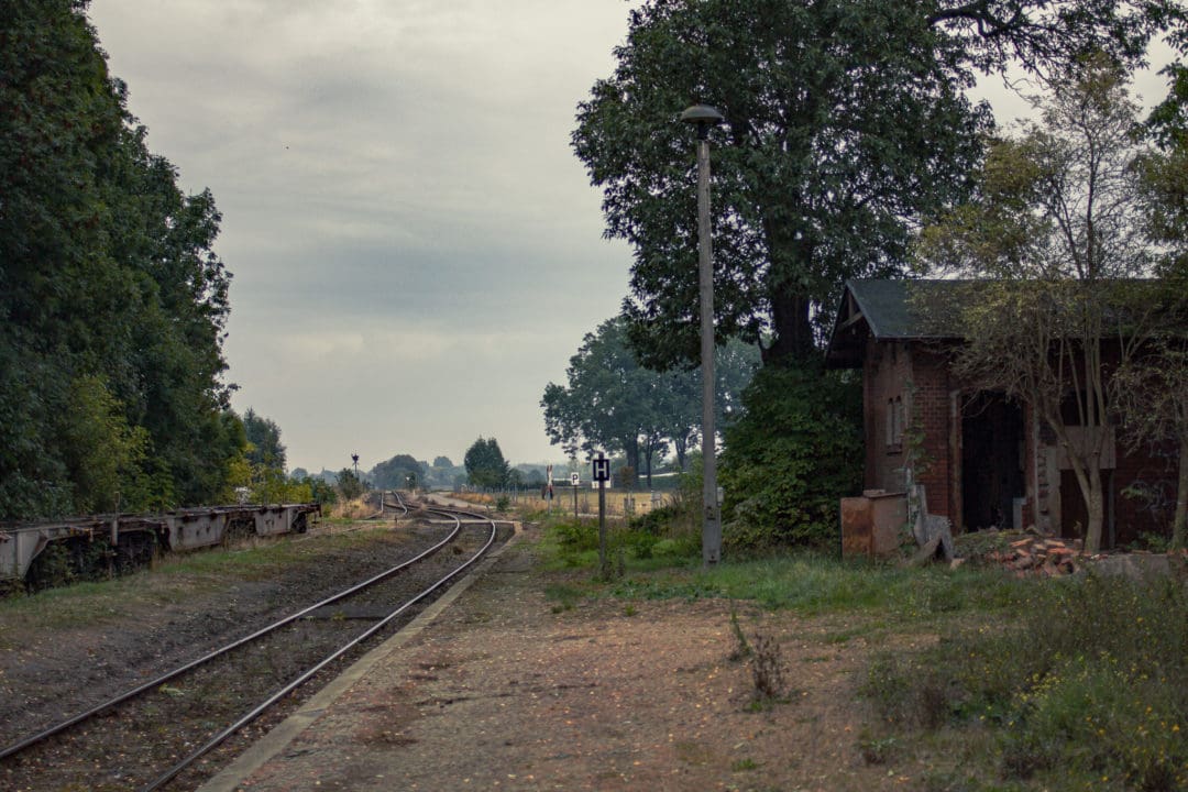 Blick über den ehemaligen Bahnsteig. Auf dem linken Gleis stehen abgestellte Waggons. Auf der rechten Seite liegen einige Ziegel vor einem heruntergekommenen Schuppen. Der Himmel ist grau, die Stimmung ist gedrückt.