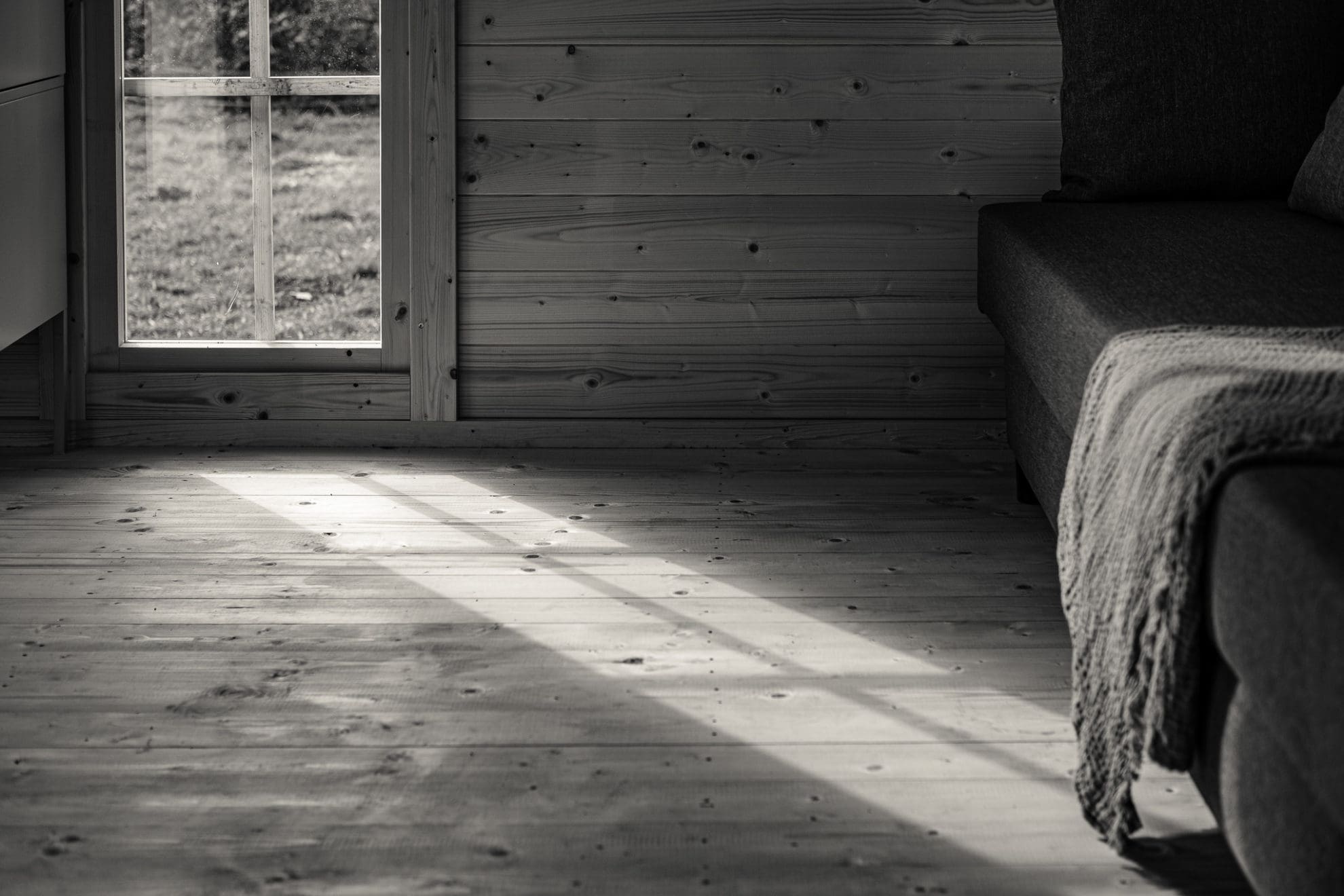 Sonnenstrahlen bilden das Fenster auf dem Holzboden ab. Der Holzboden zeigt im Schattenspiel seine raue Struktur. Im rechten Teil des Bildes fallen die Sonnenstrahlen auf eine Decke, die von einem Sofa herunterhängt.