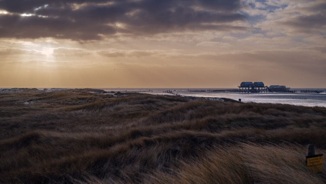 Das Gras auf den Dünen wiegt sich im Wind, die Sonne wirft Sonnenstrahlen durch die Wolkendecke und im Wasser stehen Pfahlbauten.
