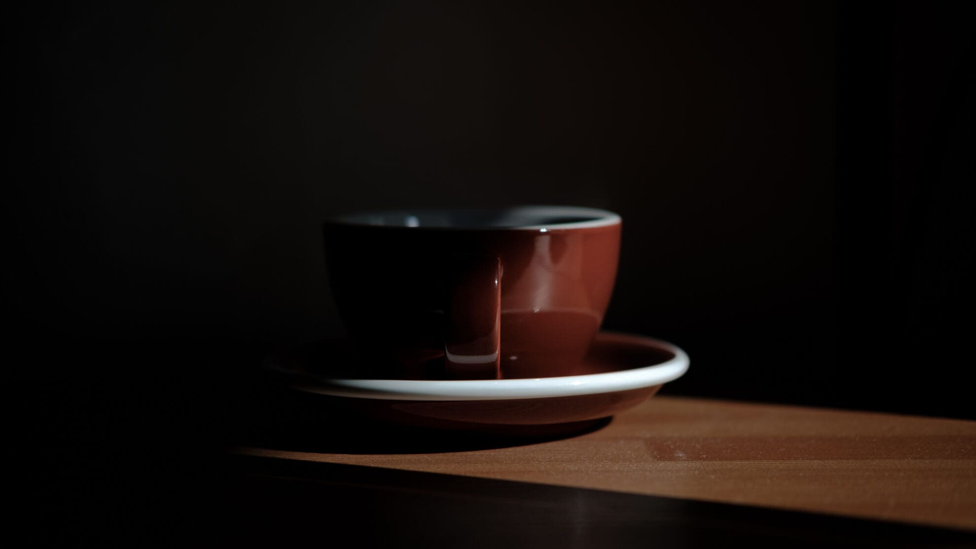 Eine braune Kaffeetasse steht mit Henkel zum Betrachter auf einer Untertasse mit weißem Rand. Die Tasse steht im Sonnenstrahl, rund herum ist es dunkel.