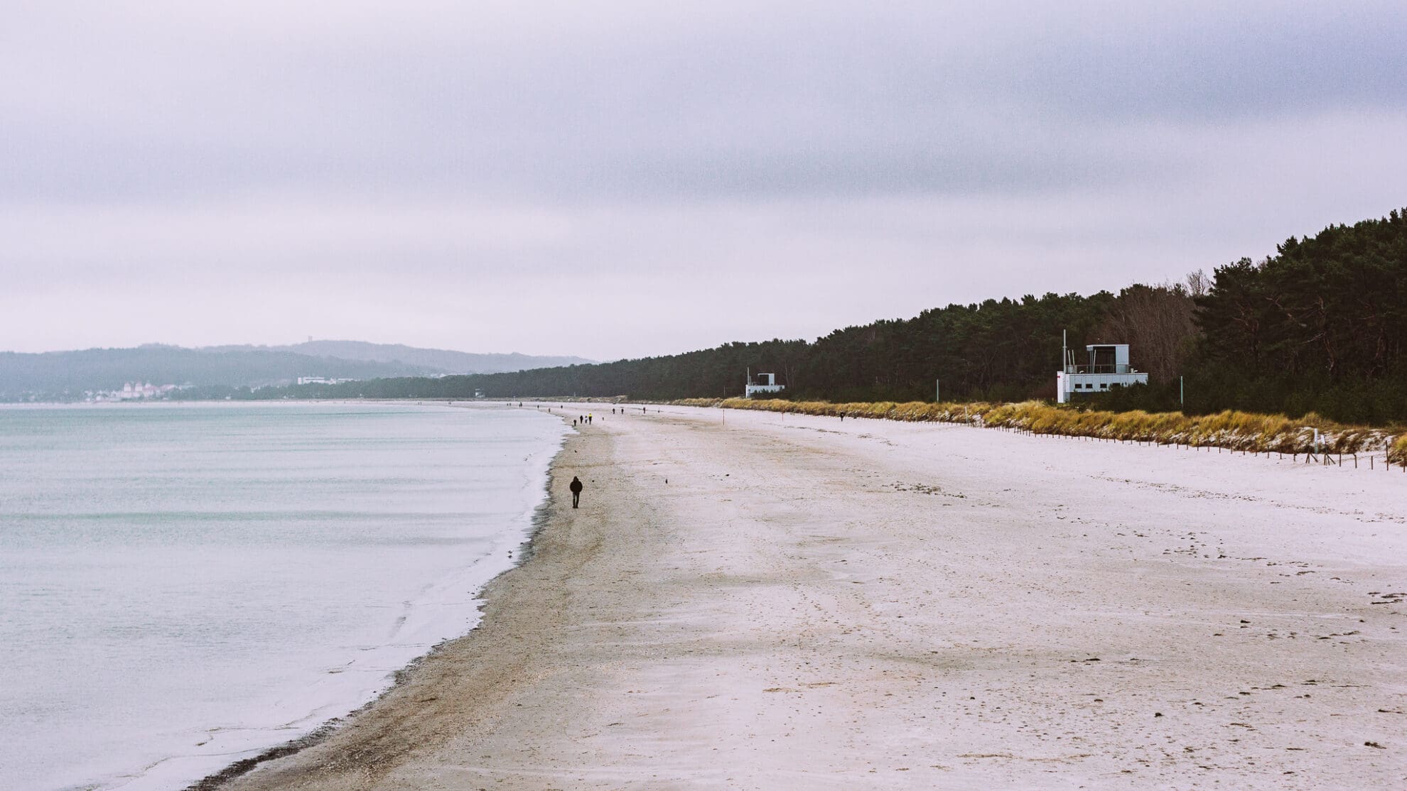 An einem weiten weißen Sandstrand läuft ein Mensch entlang, links sind der Nadelwald, Beobachtungshäuschen und Dünen zu erkennen.