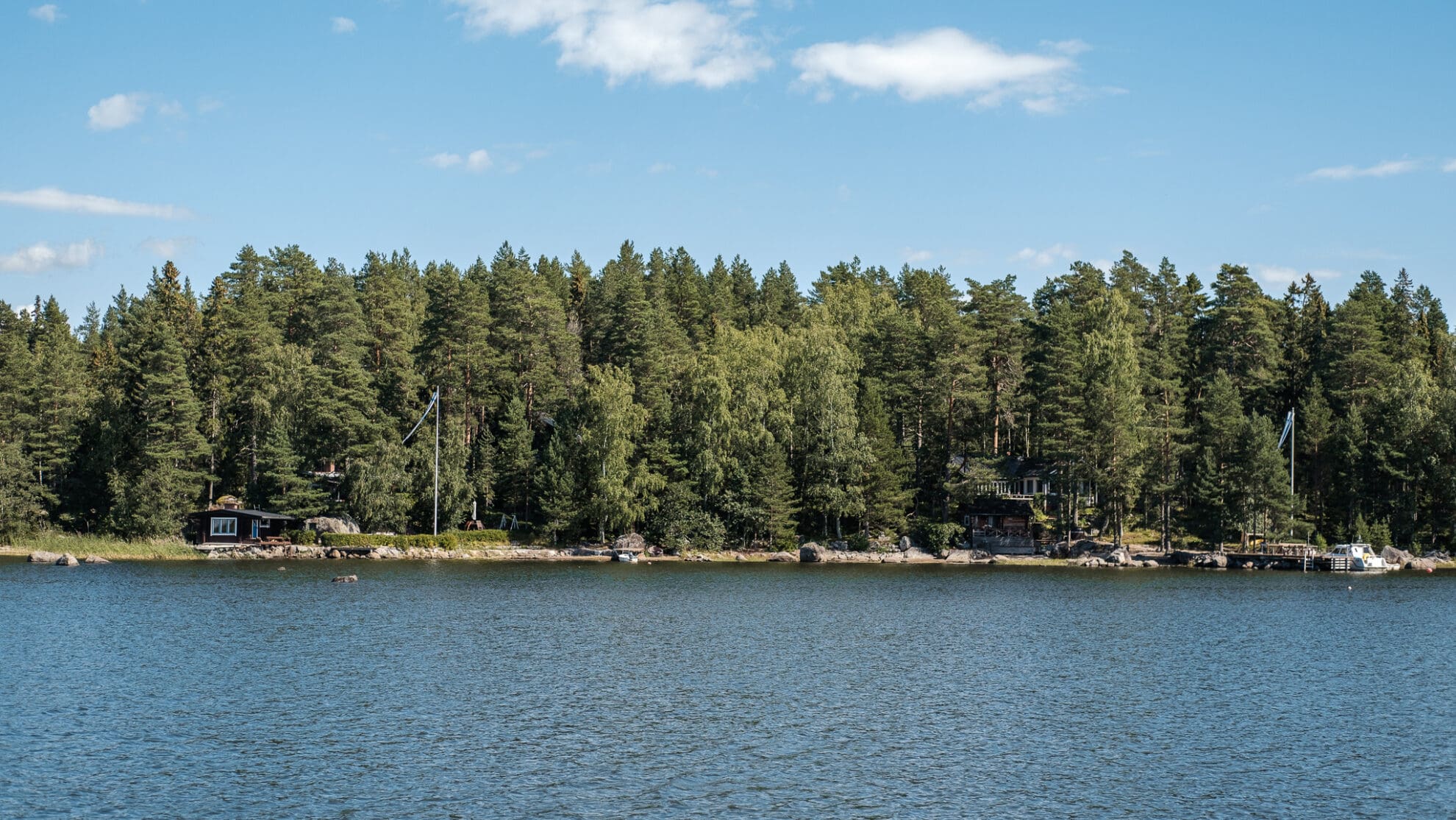 Mehrere finnische Mökkis im Wald am Ufer mit Anlegestelle und gehissten Finnlandfahnen.