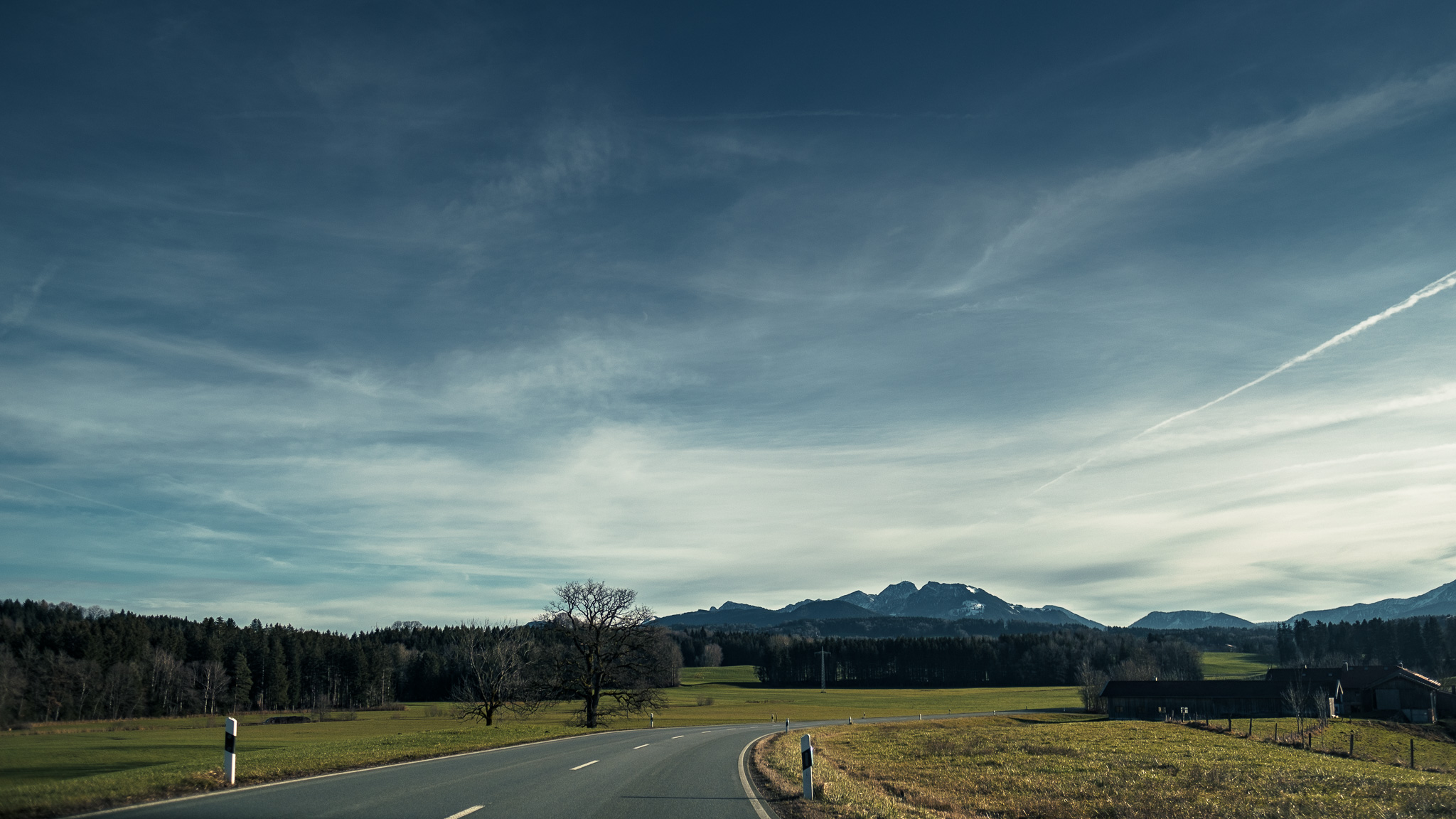 Im Vordergrund biegt eine Straße nach rechts ab, rechts und links grüne Wiesen, am Horizont erheben sich die Alpen, darüber blauer Himmel mit Schleierwolken.