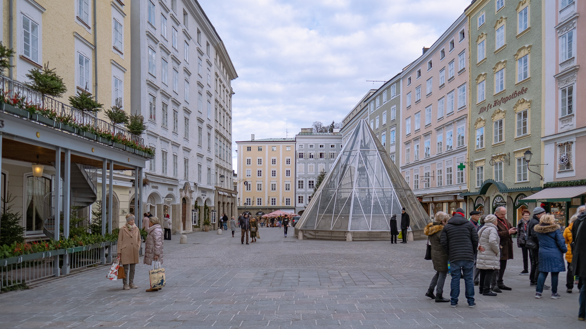 Marktplatz mit barocken Häusern rechts und links, im Vordergrund stehen Menschen, links zwei Frauen sich unterhaltend. In der Mitte des Platzes verhüllt eine gläserne Pyramide einen Springbrunnen.