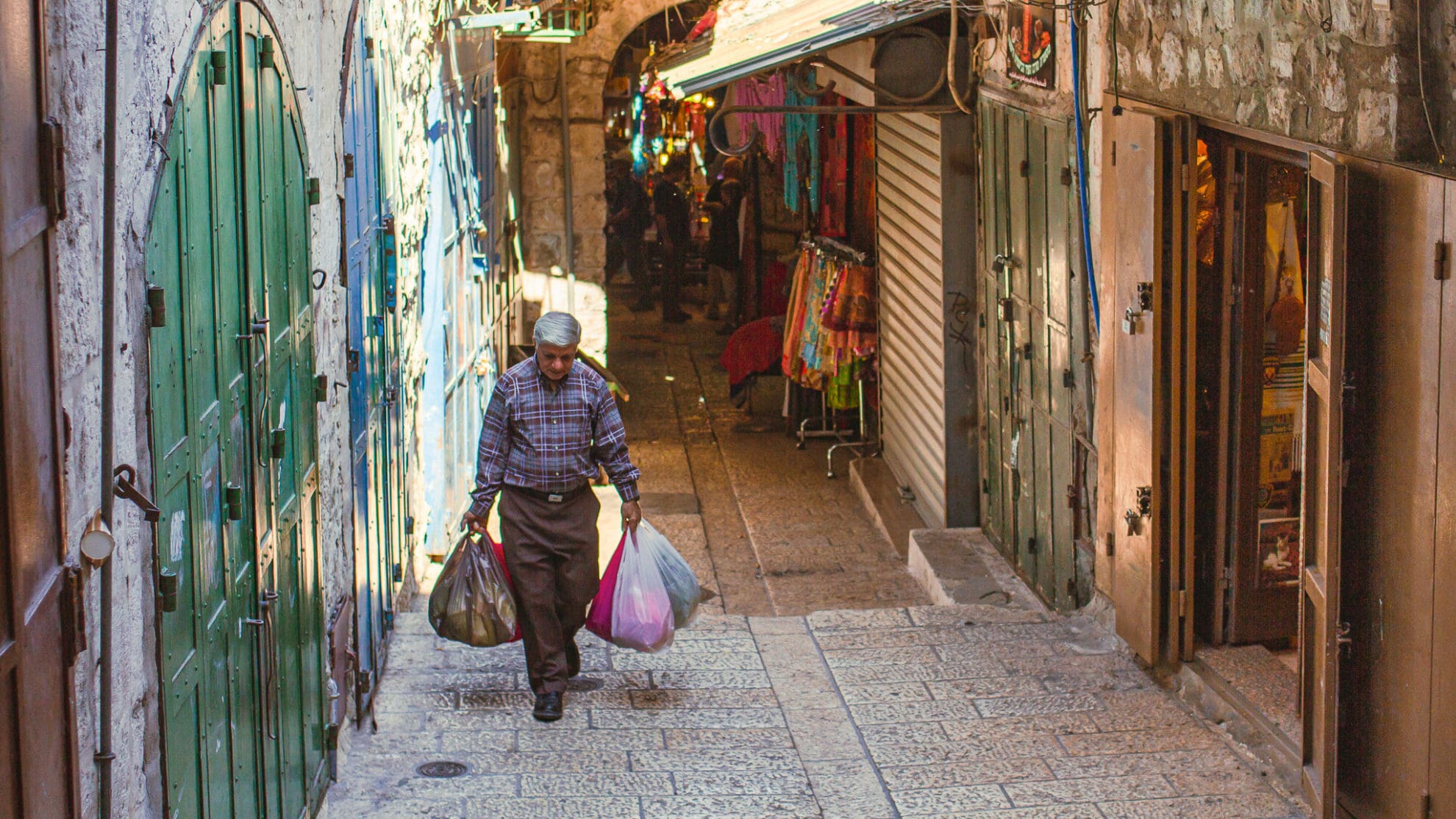 Ein Mann in einer Gasse der Jerusalemer Altstadt trägt seine Einkäufe in vielen Plastiktüten, im Hintergrund sieht man verschiedene bunte Auslagen der Geschäfte.