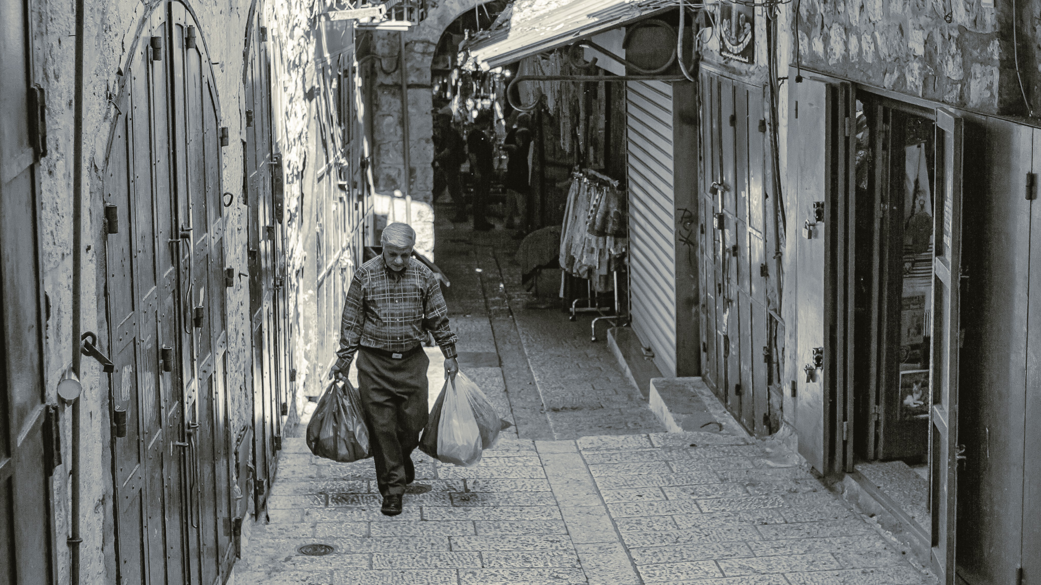 Schwarz-weiße Aufnahme eines Mannes, der in einer Gasse der Jerusalemer Altstadt seine Einkäufe in vielen Plastiktüten trägt, im Hintergrund sieht man verschiedene Auslagen der Geschäfte.