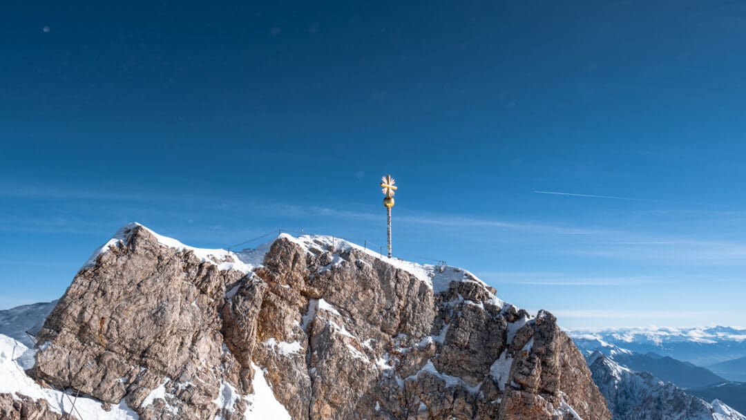 Das goldene Gipfelkreuz der Zugspitze steht auf dem Gipfel und wird von der Sonne angestrahlt, im Hintergrund sind die Alpen zu sehen, durch die Luft wirbeln einzelne Schneeflocken.
