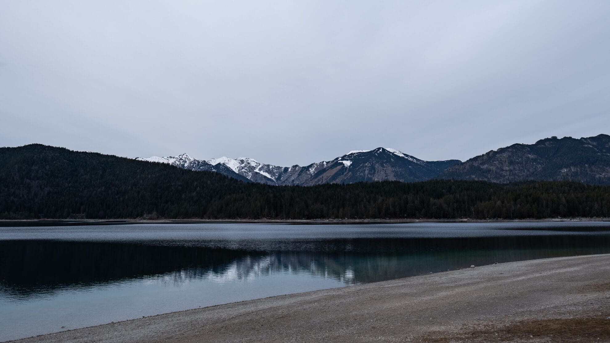 Blick über den stillen Eibsee, im Vordergrund liegt der graue Kieselstrand, im Hintergrund erheben sich mehrere schneebedeckte Gipfel vor dem grauen Himmel.