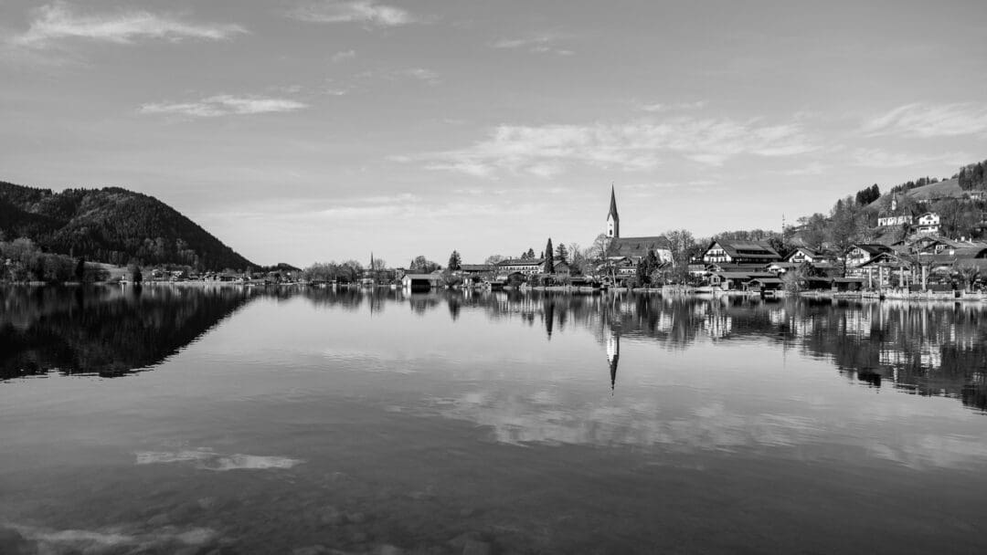 Blick auf den Ort Schliersee, im Vordergrund der See, in dem sich die Silhouette des Ortes mit dem herausstechenden Kirchturm spiegelt.