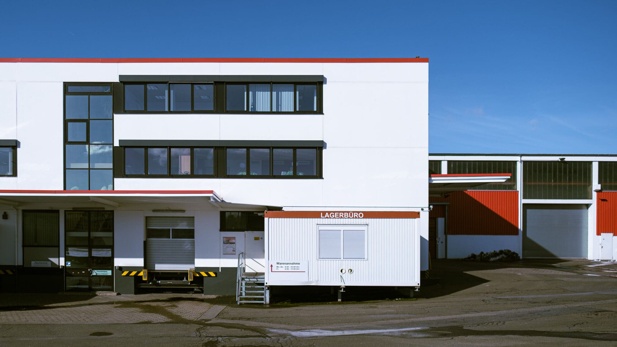 Dreistöckiges weißes Gebäude mit einem Container davor mit der Aufschrift Lagerbüro, rechts daneben eine Lagerhalle mit weiß-roter Fassade.