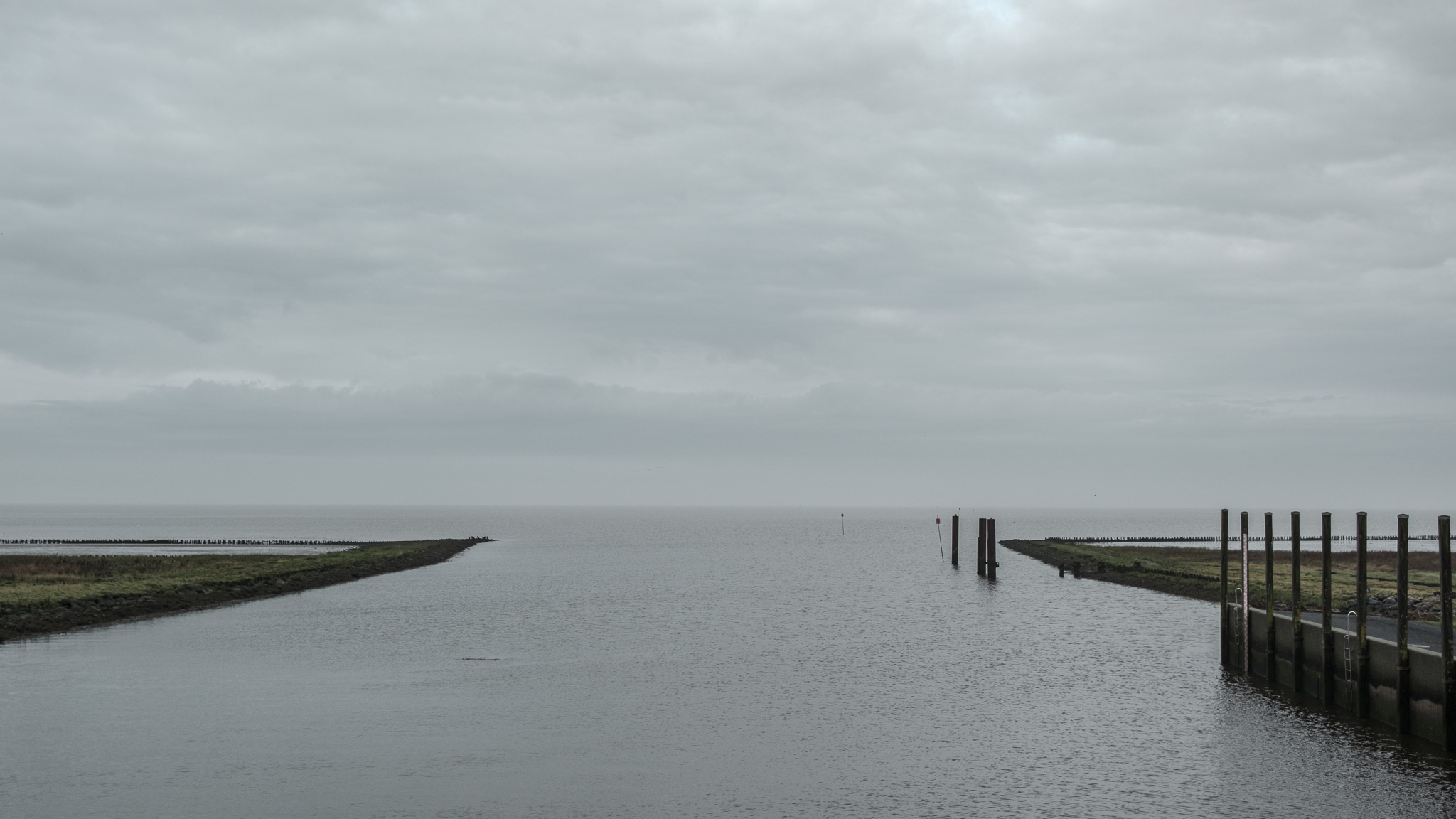 Blick von der Schleuse auf die Einfahrt auf das Wattenmeer hinaus, rechts stehen Poller, links ist der flache Uferbereich zu sehen, am Horizont der grau bedeckte Himmel.